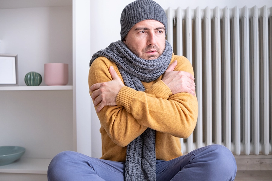 A male homeowner wearing a sweater, hat, and scarf is cold as he sits next to a malfunctioning furnace in his home in O’Fallon, IL.