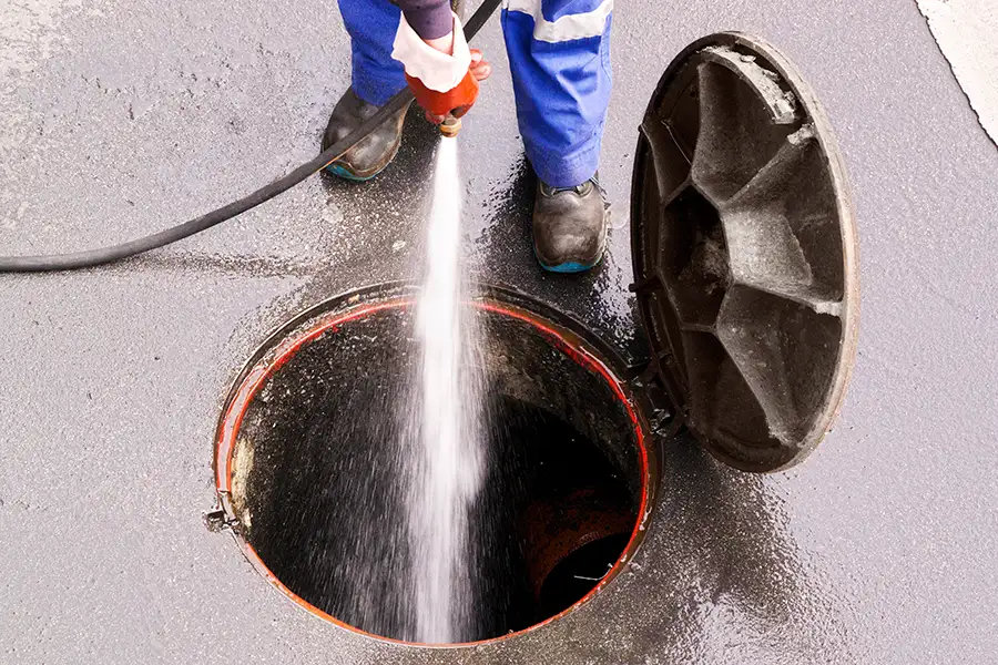 The image shows a worker standing near an open manhole, using a high-pressure water hose to clean the inside. O'Fallon, IL